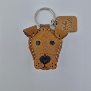 May include: A brown leather dog-shaped keychain with black eyes and nose. The keychain has a metal ring and a small tag with the text "Boho Circle". The dog's features are stitched with white thread.