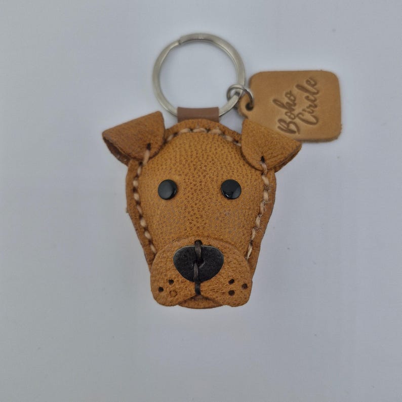 May include: A brown leather dog-shaped keychain with black eyes and nose. The keychain has a metal ring and a small tag with the text "Boho Circle". The dog's features are stitched with white thread.