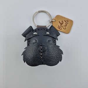 Pu&ograve; includere: Un portachiavi a forma di cane Schnauzer in pelle nera, con un portachiavi argentato e un piccolo cartellino marrone con la scritta "Boho Circle". Il muso del cane &egrave; dettagliato con cuciture e pelle strutturata.