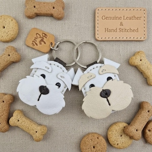Pu&ograve; includere: Due portachiavi a forma di cane in pelle bianca con dettagli beige e occhi neri, circondati da biscotti per cani. Una piccola etichetta recita "Boho Circle". Un'etichetta in pelle recita "Genuine Leather & Hand Stitched".