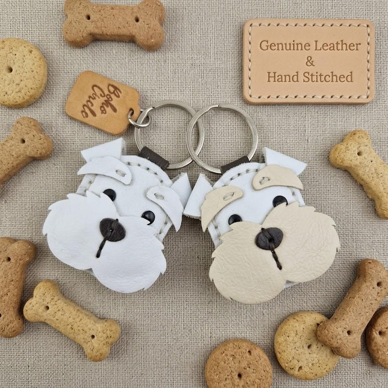 Pu&ograve; includere: Due portachiavi a forma di cane in pelle bianca con dettagli beige e occhi neri, circondati da biscotti per cani. Una piccola etichetta recita "Boho Circle". Un'etichetta in pelle recita "Genuine Leather & Hand Stitched".