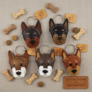 May include: Several handcrafted leather dog head keyrings in shades of brown and black, each with a metal ring. The keyrings are surrounded by dog biscuits and tags that read "Boho Circle". The image also includes a leather tag that reads "Genuine Leather & Hand Stitched."