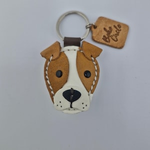 May include: A leather dog-shaped keychain with brown and white details, black eyes, and a black nose. The keychain has a silver ring and a small, brown leather tag with the words "Boho Circle" embossed on it.