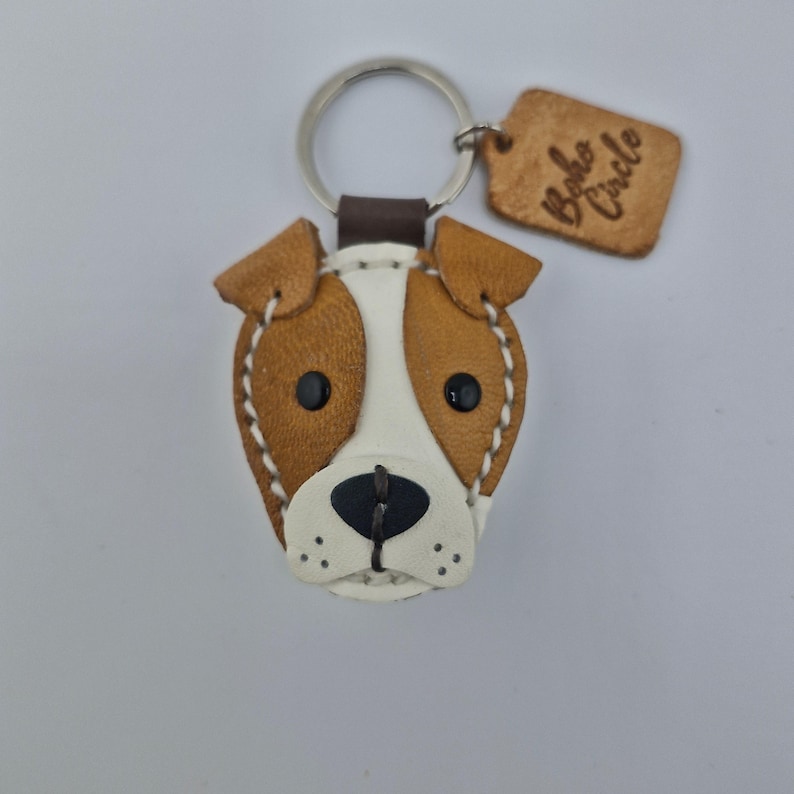 May include: A leather dog-shaped keychain with brown and white details, black eyes, and a black nose. The keychain has a silver ring and a small, brown leather tag with the words "Boho Circle" embossed on it.