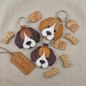 Könnte beinhalten: Handgenähte Leder-Hunde-Kopf-Schlüsselanhänger in Braun, Beige und Weiß. Jeder Schlüsselanhänger hat einen Metallring und ein kleines Lederetikett. Knochenförmige Hundekekse und ein Lederetikett mit dem Text "Genuine Leather & Hand Stitched" sind ebenfalls vorhanden.