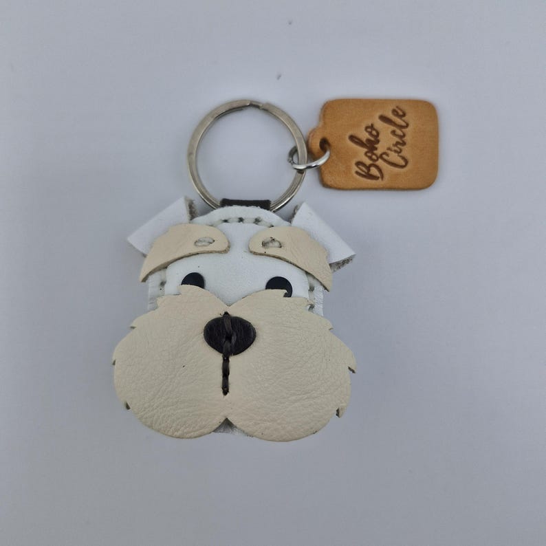 Pu&ograve; includere: Portachiavi a forma di cane in pelle bianca e beige con un portachiavi argentato. Il viso del cane &egrave; dettagliato con occhi neri e un naso nero. Un piccolo tag in pelle beige con la scritta "Boho Circle" &egrave; attaccato al portachiavi.
