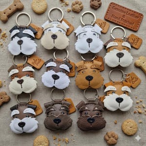 Pu&ograve; includere: Una collezione di portachiavi in pelle fatti a mano a forma di cane, in vari colori, tra cui nero, bianco, marrone e beige. Ogni portachiavi presenta un anello di metallo e una piccola etichetta. I portachiavi sono circondati da biscotti per cani e un'etichetta in pelle con la scritta "Genuine Leather & Hand Stitched".