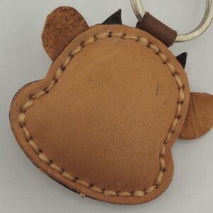 May include: A tan leather keychain in the shape of a cow's head. The keychain has a stitched border and small black horns. A silver keyring and brown leather loop are attached.