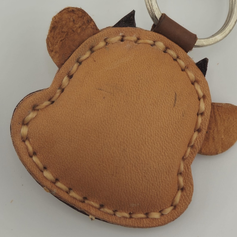 May include: A tan leather keychain in the shape of a cow's head. The keychain has a stitched border and small black horns. A silver keyring and brown leather loop are attached.