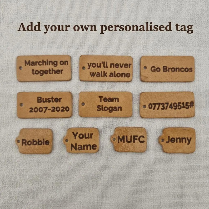 May include: A collection of rectangular, light brown tags with rounded corners. Each tag features engraved text, including phrases like "Marching on together" and names such as "Robbie" and "Jenny". The text "Add your own personalized tag" is at the top.