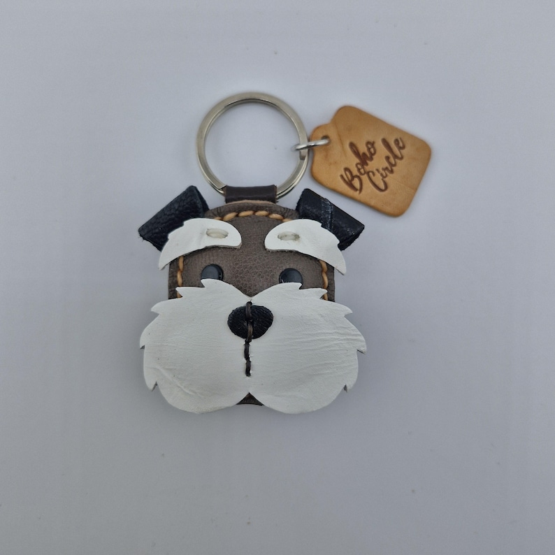 Pu&ograve; includere: Portachiavi a forma di cane in pelle con anello argentato. Il cane ha un corpo marrone, pelliccia bianca e orecchie nere. Un piccolo cartellino di legno con la scritta "Boho Circle" &egrave; attaccato.
