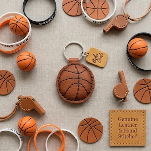 May include: A collection of basketball-themed items, including a leather keychain, whistles, bracelets, and miniature basketballs. The keychain is round and features a stitched basketball design. The bracelets are inscribed with "Court" and "Hoops". A leather tag reads "Boho Circle".