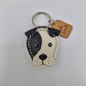 May include: A handmade leather keychain in the shape of a dog's face, featuring black and white patches, stitched details, and a metal keyring. A small, brown tag with the text "Soho Circle" is attached.