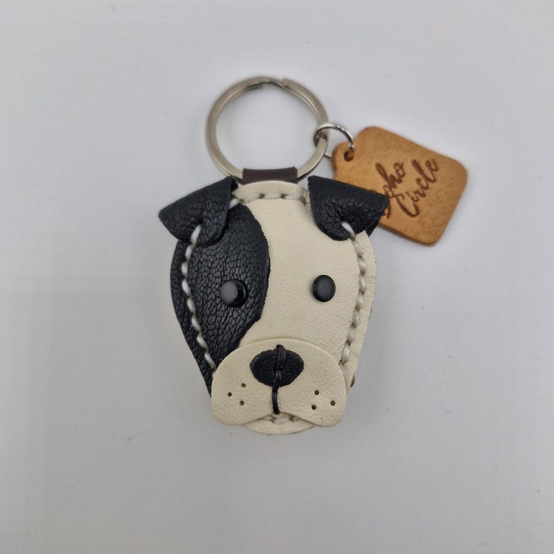 May include: A handmade leather keychain in the shape of a dog's face, featuring black and white patches, stitched details, and a metal keyring. A small, brown tag with the text "Soho Circle" is attached.