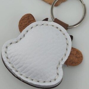 May include: A white leather keychain in the shape of a bell with brown accents and a silver key ring. The keychain has brown ears and a brown leather loop connecting it to the key ring. The edges are stitched with white thread.