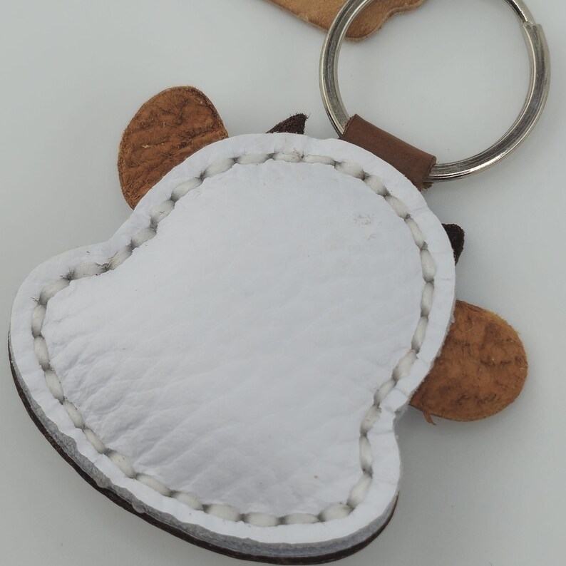 May include: A white leather keychain in the shape of a bell with brown accents and a silver key ring. The keychain has brown ears and a brown leather loop connecting it to the key ring. The edges are stitched with white thread.