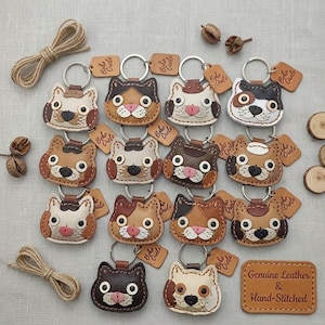 May include: A collection of cat-shaped leather keychains in various colors including brown, black, white, and tan. Each keychain features a metal ring and a small rectangular tag embossed with the text Boho Circle. Some tags read Genuine Leather & Hand-Stitched.