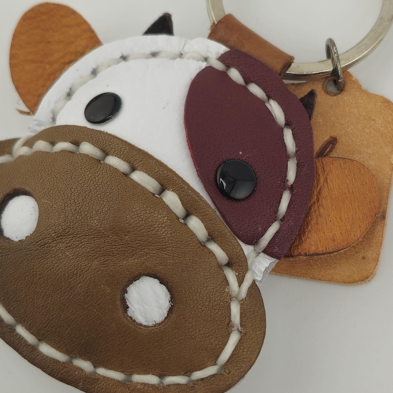 May include: A handcrafted leather keychain in the shape of a cow's face. The keychain features brown, white, and burgundy leather pieces stitched together with white thread. The cow has black button eyes and a small leather tag.
