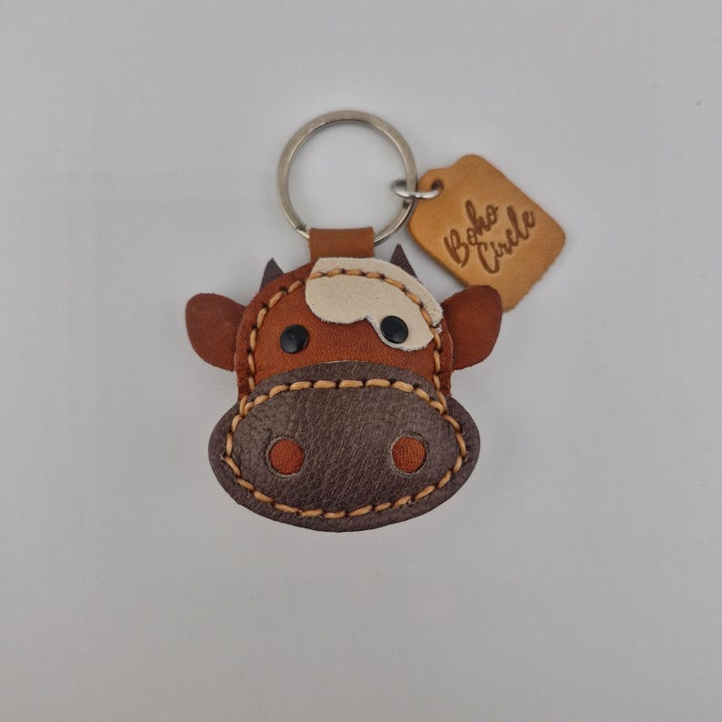 May include: A brown and tan leather cow keychain with a silver keyring. The cow has black eyes and a white patch on its forehead. A small, brown leather tag with the words "Boho Circle" is attached to the keyring.