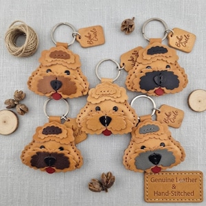 Könnte beinhalten: Fünf Schlüsselanhänger aus Leder in Form von Hundeköpfen mit lockiger Fellstruktur, Metallringen und kleinen rechteckigen Anhängern mit der Prägung Boho Circle. Jedes Stück hat eine individuell gefärbte Nase und Zunge in Schwarz, Braun oder Grau.
