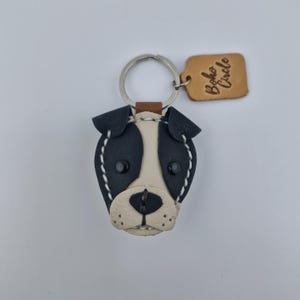 May include: A handmade leather dog keychain with a black and white face, stitched with white thread. The keychain has a silver ring and a small brown leather tag that says "Boho Circle".