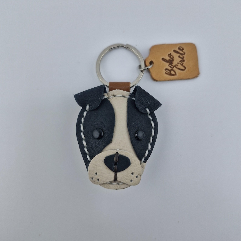 May include: A handmade leather dog keychain with a black and white face, stitched with white thread. The keychain has a silver ring and a small brown leather tag that says "Boho Circle".