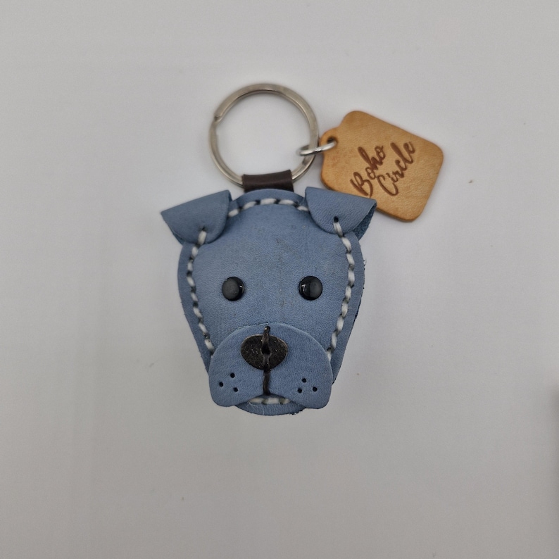 May include: A blue leather dog-shaped keychain with black eyes and a stitched outline. The keychain has a silver ring and a small wooden tag that reads "Boho Circle".