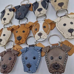 May include: A collection of handmade leather dog keychains in various colors, including brown, blue, and cream. Each keychain features a detailed dog face design with stitched accents and a metal keyring.