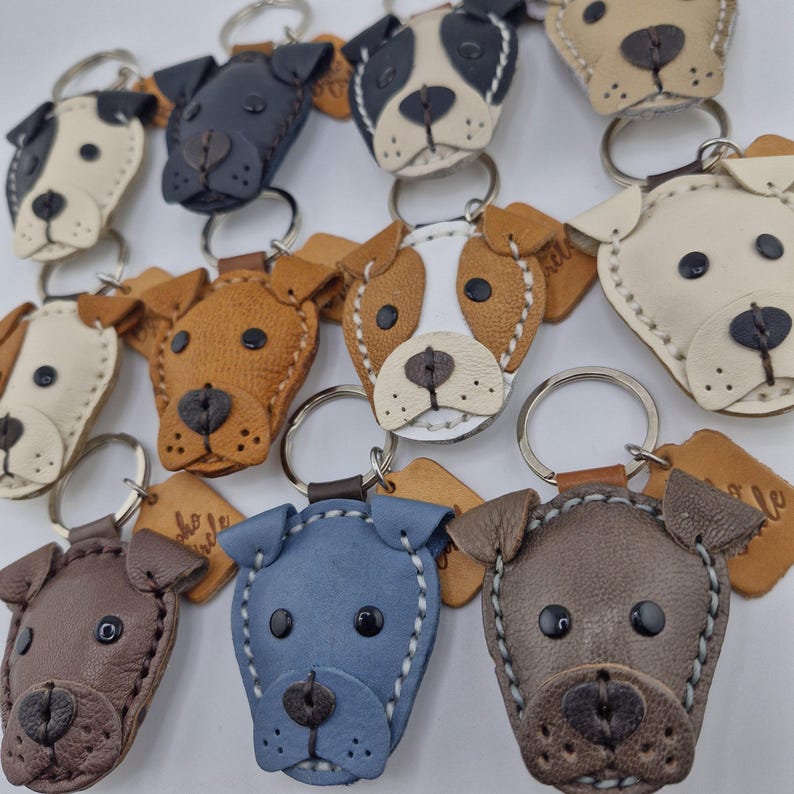 May include: A collection of handmade leather dog keychains in various colors, including brown, blue, and cream. Each keychain features a detailed dog face design with stitched accents and a metal keyring.