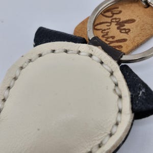 May include: A handmade leather keychain featuring a panda bear design. The keychain has a white face, black ears, and a stitched border. A small wooden tag with the text "Boho Circle" is attached.