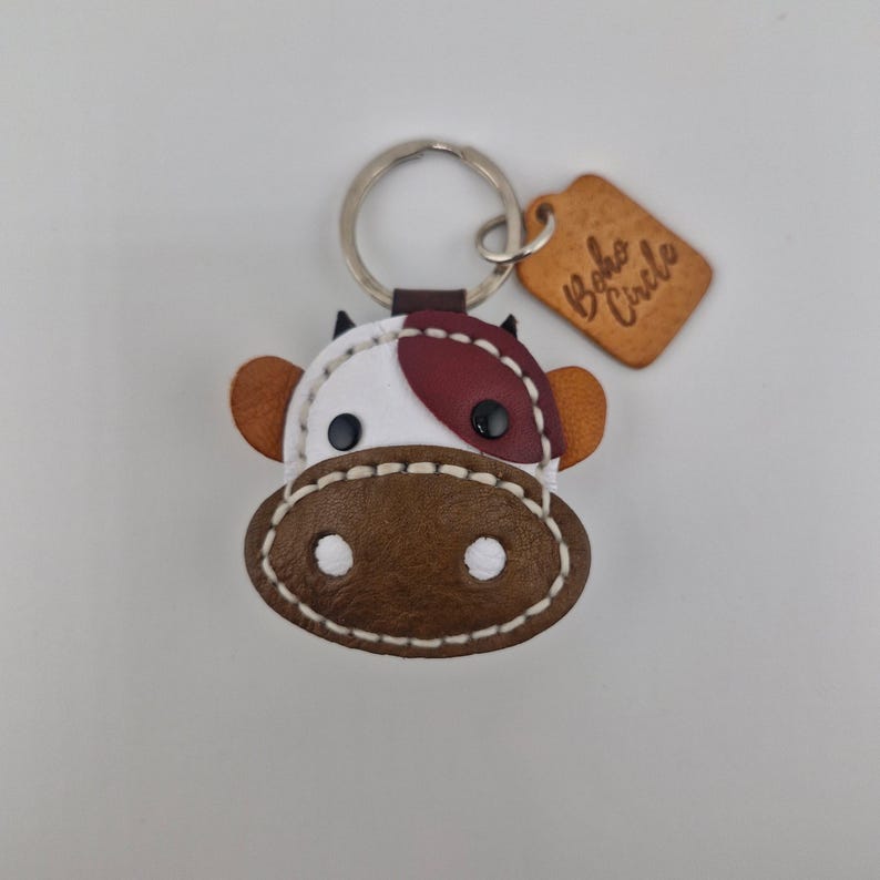 May include: A cow-shaped leather keychain with a silver keyring. The cow has a white face with a maroon patch, brown ears, and a brown muzzle. The keychain includes a small, tan tag with the text "Boho Circle".