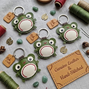 May include: Three round frog keychains made of green and cream leather with red stitching, each featuring a metal ring and a small tan leather tag embossed with the text Boho Circle. A rectangular leather label reads Genuine Leather & Hand-Stitched.