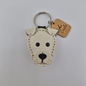 May include: A cream-colored leather dog keychain with black eyes and nose. The keychain has a silver ring and a small, brown tag that reads "Soho Circle". The dog's features are stitched with cream-colored thread.