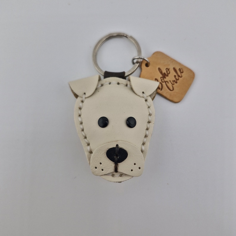 May include: A cream-colored leather dog keychain with black eyes and nose. The keychain has a silver ring and a small, brown tag that reads "Soho Circle". The dog's features are stitched with cream-colored thread.