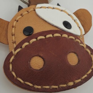 May include: A handmade leather keychain in the shape of a cow's face. The keychain features a brown snout with two circular nostrils, black eyes, and a white patch on the forehead. The edges are stitched with a light tan thread.