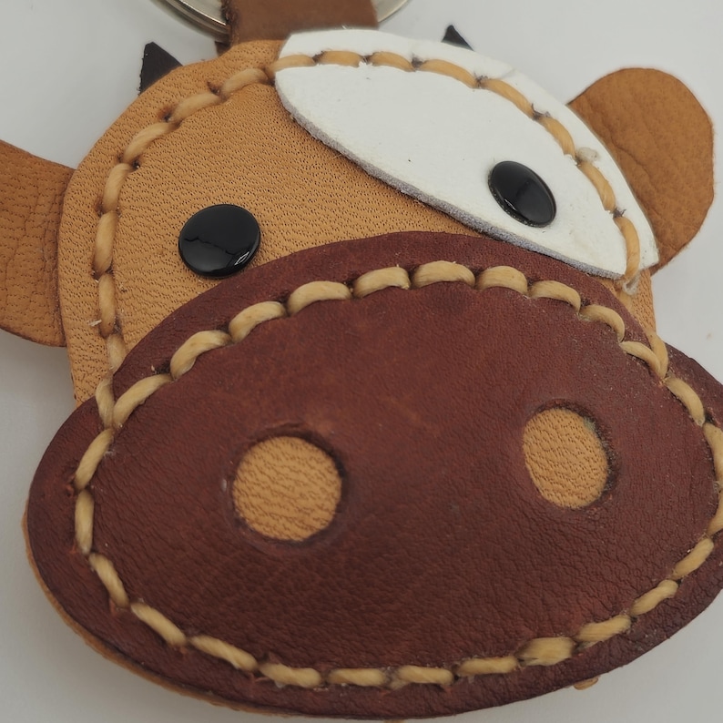 May include: A handmade leather keychain in the shape of a cow's face. The keychain features a brown snout with two circular nostrils, black eyes, and a white patch on the forehead. The edges are stitched with a light tan thread.