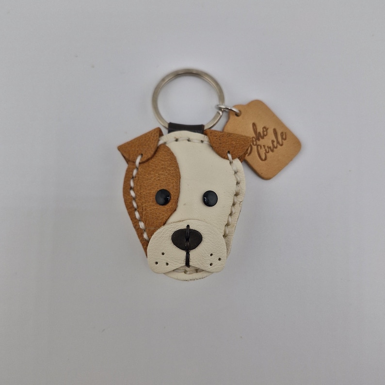 May include: A handmade leather dog-shaped keychain. The keychain features a brown and white design with black button eyes and a stitched outline. A small, brown tag with the text "Soho Circle" is attached to the keyring.