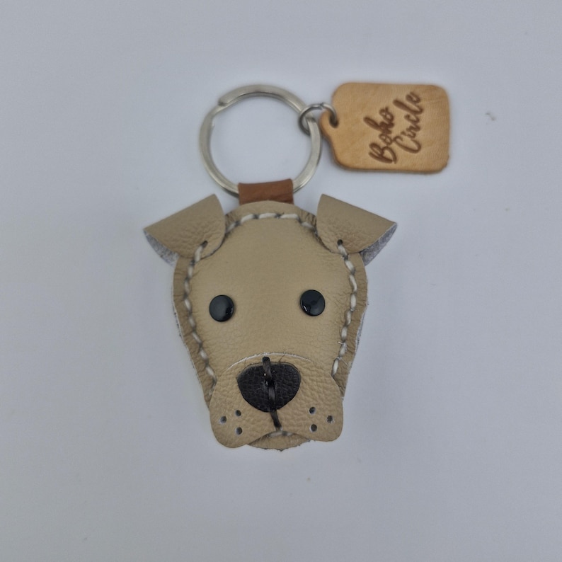 May include: Tan leather dog-shaped keychain with black button eyes and a stitched outline. The keychain has a silver ring and a small leather tag with the text "Boho Circle".