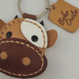 May include: A handcrafted leather keychain in the shape of a cow's face. The keychain features a brown snout, tan face, white patch, and black eyes. The words "Boho Circle" are embossed on a small leather tag.