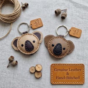 Könnte beinhalten: Zwei koalaförmige Schlüsselanhänger aus Leder mit Metallringen und kleinen beigen Anhängern mit der Prägung Boho Circle. Ein Anhänger ist hellbeige, der andere mittelbraun. Ein rechteckiges Lederschild trägt die Aufschrift Genuine Leather & Hand-Stitched.
