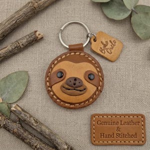 May include: A leather sloth keyring with a metal ring and a small tag that says "Boho Circle". The sloth face is brown with stitched details and black eyes. The keyring is made of brown leather and has hand-stitched details. The tag says "Boho Circle".