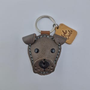 May include: A brown leather dog-shaped keychain with black button eyes and a stitched outline. The keychain has a silver ring and a small leather tag with the text "Boho Circle".