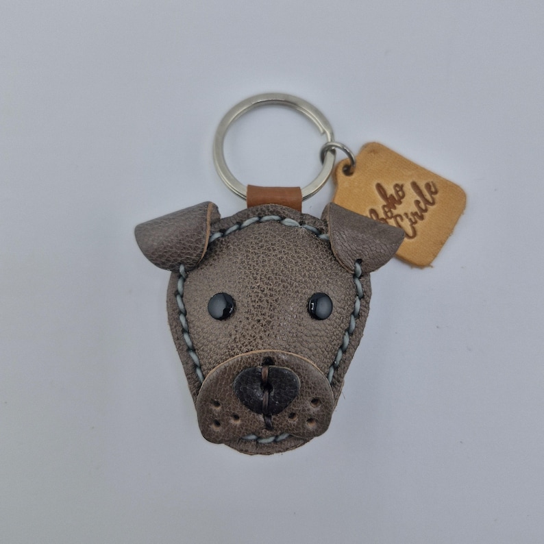 May include: A brown leather dog-shaped keychain with black button eyes and a stitched outline. The keychain has a silver ring and a small leather tag with the text "Boho Circle".