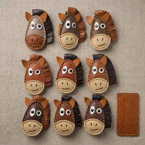 Puede incluir: Nueve llaveros de cuero con forma de caballo en varios tonos de marrón, cada uno con un hocico claro, ojos blancos con pupilas negras y una crin con flecos. Una etiqueta de cuero rectangular dice: GENUINE LEATHER & HAND STITCHED.