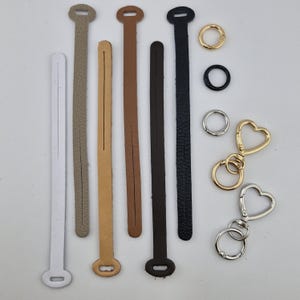 Può includere: Una collezione di sei cinturini in pelle lunghi e sottili nei colori bianco, beige, marrone chiaro, marrone, marrone scuro e nero, ciascuno con un'estremità fessurata. Sono inclusi due anelli metallici semplici in oro e nero e due portachiavi a forma di cuore in oro e argento.
