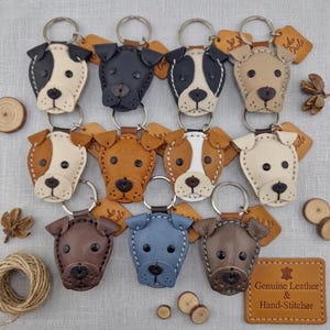 May include: A collection of eleven leather dog-shaped keychains in various colours including white, black, tan, brown, and blue. Each keychain features stitched edges, dark eyes, and a nose, attached to a metal ring with a small leather tag.
