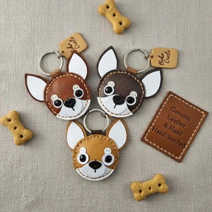 May include: Three leather Chihuahua keychains in shades of brown, tan, and white, with black button eyes and a stitched design. Each keychain has a metal ring and a small tag. The image also includes dog biscuits and a leather tag that says "Genuine Leather & Hand Stitched".