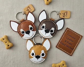 May include: Three leather Chihuahua keychains in shades of brown, tan, and white, with black button eyes and a stitched design. Each keychain has a metal ring and a small tag. The image also includes dog biscuits and a leather tag that says "Genuine Leather & Hand Stitched".