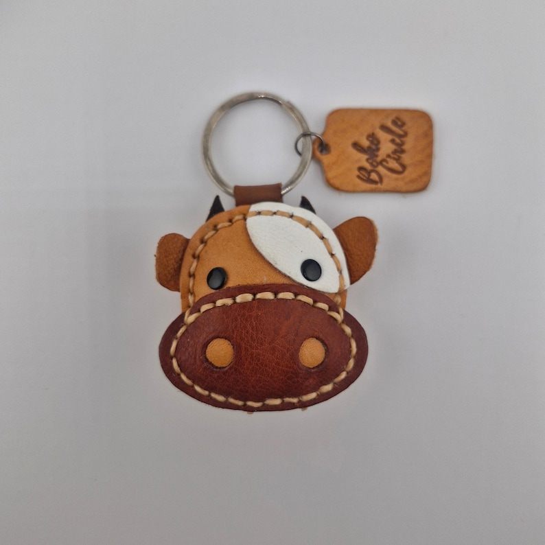 May include: A handcrafted leather cow keychain with a metal ring. The cow's face is brown, white, and tan, with stitched details and black eyes. A small leather tag with the text "Boho Circle" is attached.