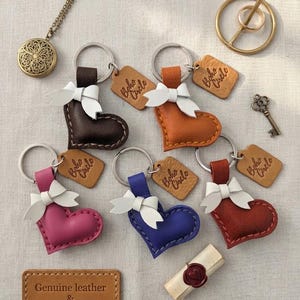 May include: Five heart-shaped leather keychains in dark brown, orange, pink, blue, and red, each with a white bow, a metal ring, and a tan tag reading Boho Circle. Includes a gold locket, a gold ring, a small key, and a wax-sealed scroll.